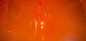 Fondo oscuro con iluminación naranja intensa. Puntos y trazos de luz blancos y rojos dispersos. Una estructura rectangular oscura se observa en el lado izquierdo