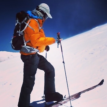 Aron Ralston, visto de lado, subiendo una ladera nevada con esquís y bastones. Viste chaqueta naranja, pantalón oscuro, gorra y mochila bajo un cielo azul