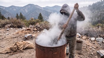 Un hombre con capucha y rostro cubierto sostiene un palo dentro de un barril metálico del que sale humo blanco, en un terreno montañoso.