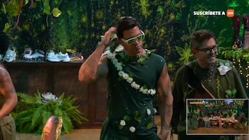 Un hombre con traje verde y gafas de sol se toca la cabeza junto a otro hombre en un escenario decorado con plantas