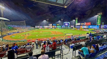 Vista aérea de un campo de béisbol iluminado por focos, con jugadores en el terreno y gradas llenas de espectadores bajo un cielo nocturno.