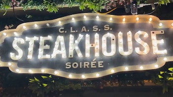 Un letrero negro con borde de luces y letras iluminadas que dicen C. L. A. S. S. Steakhouse Soirée, rodeado por plantas verdes