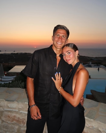 Un hombre y una mujer sonrientes se abrazan al atardecer. Él viste camisa negra y ella un vestido negro, con el mar y una piscina de fondo
