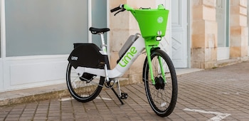 Bicicleta eléctrica Lime de colores blanco, negro y verde brillante, con cesto delantero, asiento, pedales, batería y caballete, sobre suelo adoquinado