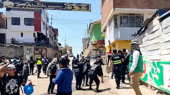 Policías con equipos de protección y ciudadanos en una calle inclinada frente a viviendas y un letrero que dice El Almacén popular.