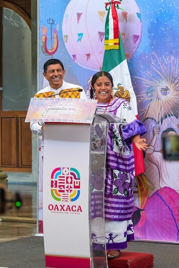 Gobernador Salomón Jara Cruz sonríe junto a mujer oaxaqueña en traje tradicional morado y blanco, detrás de atril con logo de Oaxaca y Guelaguetza 2026