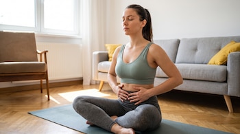 Mujer sentada con piernas cruzadas en colchoneta de yoga, ojos cerrados, manos sobre su abdomen, en una habitación con sofá y silla.