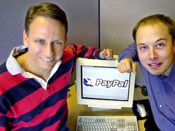 Dos hombres sonrientes, Peter Thiel con un polo a rayas y Elon Musk con una camisa morada, posan junto a un monitor que muestra el logo de PayPal