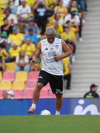 Un jugador de fútbol con camiseta blanca sin mangas y pantalón corto negro maneja la pelota con el pie en la cancha