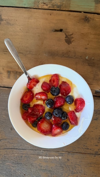 Un plato blanco con yogur, fresas cortadas, arándanos y miel, con una cuchara de metal, sobre una mesa de madera. Incluye el texto: Mi desayuno by isi