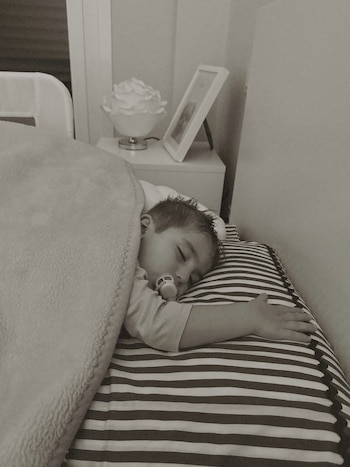 Un niño pequeño duerme con un chupete. Está cubierto por una manta y recostado en una almohada a rayas. Se ven una mesita de noche, una lámpara y un cuadro