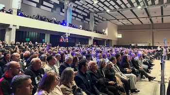 Gran salón con público sentado en el nivel inferior y de pie en un balcón. Se observan banners colgados, luces y equipo de cámara en un escenario lateral