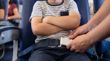 Una persona adulta abrocha el cinturón de seguridad de un niño con camiseta de rayas y pantalón oscuro, sentado en un asiento azul de avión.
