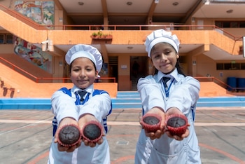 Dos niñas con uniforme escolar y gorro blanco sostienen cuatro magdalenas de chocolate hacia la cámara en el patio de un colegio
