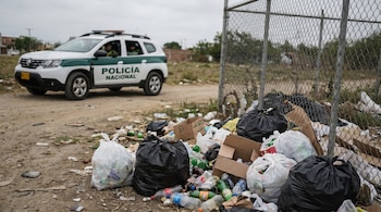 Un lote baldío lleno de bolsas de basura negras y blancas, botellas plásticas y cajas de cartón; al fondo se ve una patrulla de la Policía Nacional de Colombia.