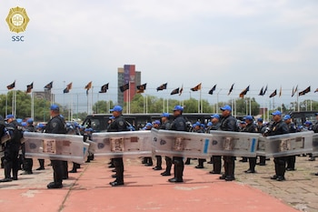 Vista lateral de un grupo de policías uniformados con cascos azules y escudos antidisturbios en formación. Al fondo se ven autobuses y edificios bajo un cielo nublado