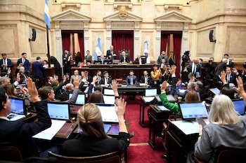 Una sala de asambleas con banderas argentinas. Múltiples personas sentadas en escritorios con computadoras levantan las manos para votar. Varios hombres en un estrado principal