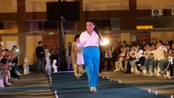Una mujer en camisa blanca y pantalón azul desfila en una pasarela azul. Hay otras modelos detrás y público sentado a ambos lados del recorrido