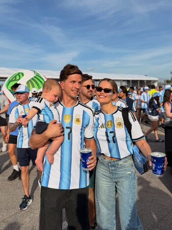 Un hombre sostiene a un bebé mientras camina junto a una mujer sonriente con gafas de sol, todos con camisetas de Argentina, entre una multitud y bajo el sol