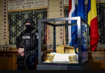 Un casco de oro y brazaletes dorados se exhiben en una vitrina de cristal. Un agente de policía con uniforme oscuro vigila. Banderas de la UE y Rumania al fondo