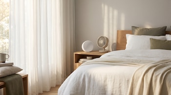 Dormitorio minimalista con cama de sábanas blancas, máquina de ruido blanco y ventilador sobre mesa de noche, junto a ventana con cortinas translúcidas.