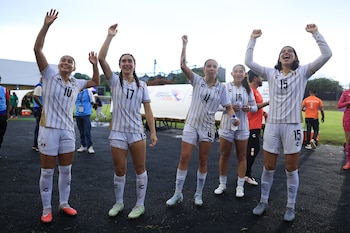 Cinco mujeres futbolistas con uniformes de México (blanco y oro) con números 10, 17, 4, 16, 15, levantan los brazos. Hay personas y un cartel