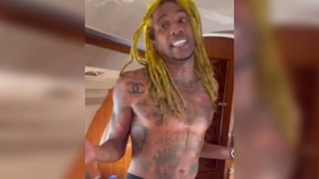 Un hombre sin camisa con rastas teñidas de amarillo y tatuajes en el torso habla en un espacio interior