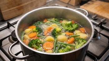 Una olla de acero inoxidable llena de brócoli, zanahorias en rodajas, patatas y judías verdes hirviendo vigorosamente sobre una estufa de gas.