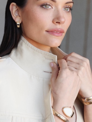 Primer plano de Eva De Dominici ajustando el cuello de su chaqueta beige, luciendo pendientes, reloj y pulseras doradas. Tiene ojos verdes