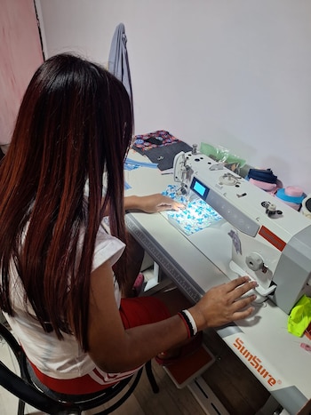 Persona con cabello castaño oscuro, camiseta blanca y shorts rojos sentada de espaldas, cosiendo una tela azul estampada en una máquina SunSure sobre una mesa blanca
