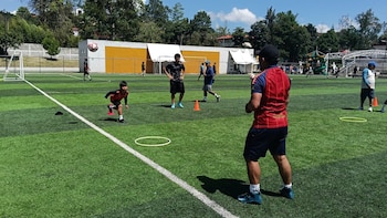 Cancha de césped artificial. Un niño con balón de fútbol en el aire. Varios hombres en el campo. Portería, conos y aros están distribuidos