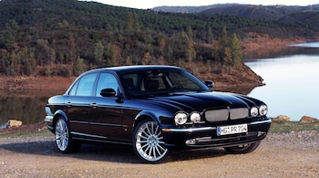 Un sedán Jaguar X-Type negro brillante está estacionado en un camino de tierra, con un lago sereno a su derecha y colinas densamente arboladas al fondo.