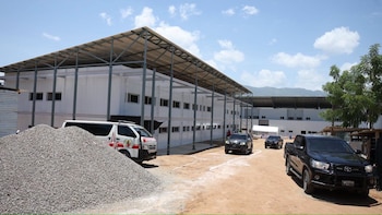 Edificio blanco modular, techo metálico, pila de grava, furgoneta blanca, todoterreno negro, camioneta pickup negra en estacionamiento de tierra