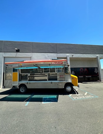 Un camión de comida de color gris y amarillo está estacionado en un aparcamiento frente a un edificio con el letrero Food Trucks