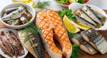 Un surtido de pescado en tabla de madera: salmón, caballa, sardinas en aceite, anchoas, con limón y hierbas. Un vaso de aceite y ensalada al fondo.