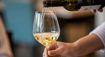 Una persona sirve vino blanco de una botella oscura en una copa de cristal. El líquido amarillo claro forma burbujas al caer en la copa.