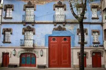 Fachada de edificio antiguo con paredes azules y descascaradas, ocho ventanas blancas, dos balcones con barandales de hierro y tres puertas dobles rojas, un árbol