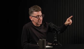 Hombre de mediana edad con gafas, Dr. Marc Brackett, sentado en una mesa con micrófono y taza, señalando con el dedo índice hacia la derecha