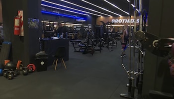 Gimnasio moderno con luces azules, piso oscuro, varias máquinas de ejercicio y pesas. Un hombre en un mostrador; una mujer levanta mancuernas en segundo plano