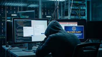 Persona con capucha sentada de espaldas frente a múltiples monitores. Las pantallas muestran código, inicio de sesión de Microsoft 365, alerta de phishing y alerta cibernética del FBI.