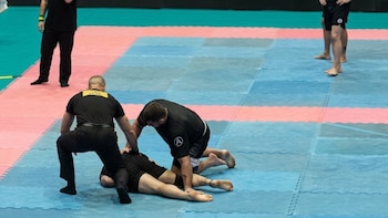Un árbitro y dos competidores de jiu-jitsu vestida con ropa negra sobre un tatami de baldosas azules y rosadas