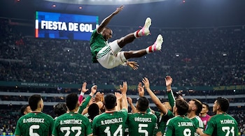 Varios futbolistas con camisetas verdes lanzan a un compañero hacia el aire en un estadio iluminado con público en las tribunas.
