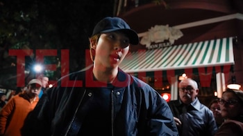 Un hombre joven con gorra y chaqueta oscura camina de noche frente a un restaurante con toldo a rayas rodeado de transeúntes