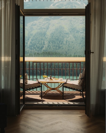 Interior de habitación con puertas abiertas hacia un balcón. En el balcón: mesa redonda, dos sillas y desayuno. Fondo: lago y montañas verdes