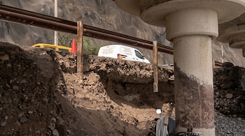 Un terreno erosionado bajo una barrera de metal oxidada, una columna de hormigón, una motocicleta estacionada y una furgoneta blanca al fondo.