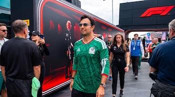 Un hombre con camiseta verde de México y gafas de sol camina entre gente en un evento. Detrás, una estructura negra con luces rojas y el logo de Fórmula 1.