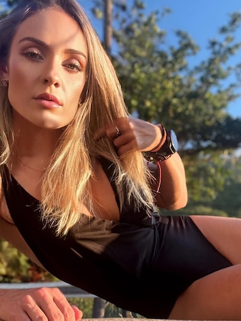 Una mujer de cabello rubio, con traje de baño negro y reloj deportivo en la muñeca, posa reclinada con vegetación y cielo azul de fondo