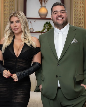Wanda Nara, con cabello rubio y vestido negro escotado con guantes largos, posa junto a Darío Barassi, quien lleva un traje verde oliva y camisa blanca