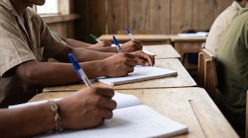 Manos y brazos de estudiantes escribiendo con bolígrafos en cuadernos sobre pupitres de madera clara en un aula escolar.