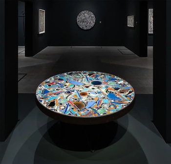 Una mesa circular de mosaico con azulejos de colores en primer plano frente a una sala de museo con paredes oscuras y obras expuestas